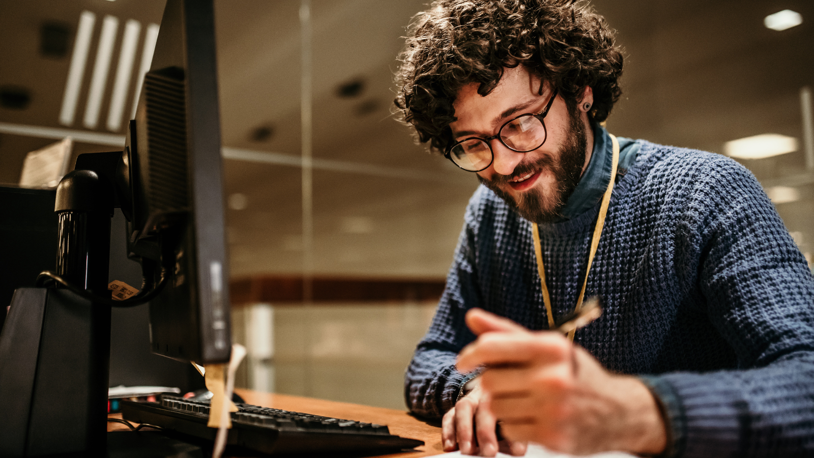 A young caucasian man with thick brown curly hair and a brown beard is wearing glasses and a comfy blue sweater while taking notes on a notepad sitting at a desk in front of a computer.